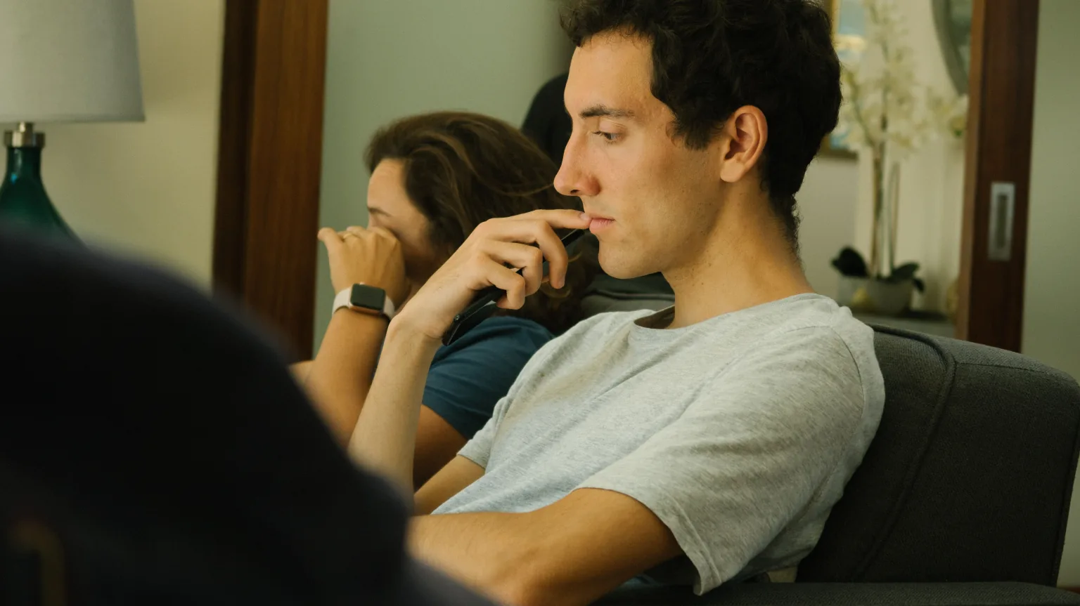 A young man sits back on a sofa holding a phone against his chin, looking away in thought, while behind him a woman rests her forehead against her hand.